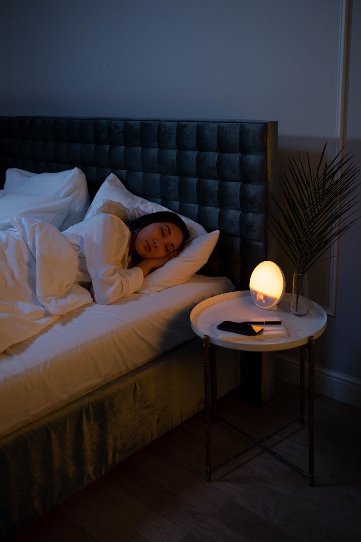 Kvinde der sover i en seng med hvidt sengetøj, med en tændt natlampe på bordet ved siden af